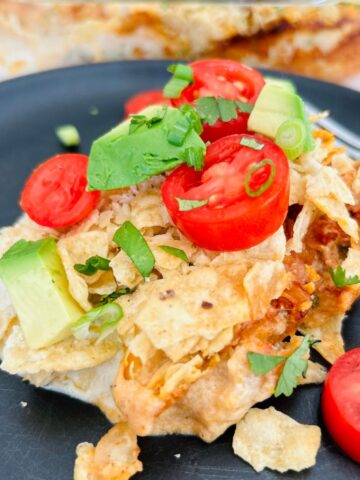 Taco casserole with tortilla chips on a black plate with sliced tomatoes, avocado, and cilantro.