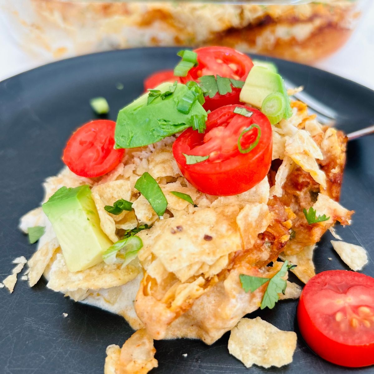 Taco casserole with tortilla chips on a black plate with sliced tomatoes, avocado, and cilantro.