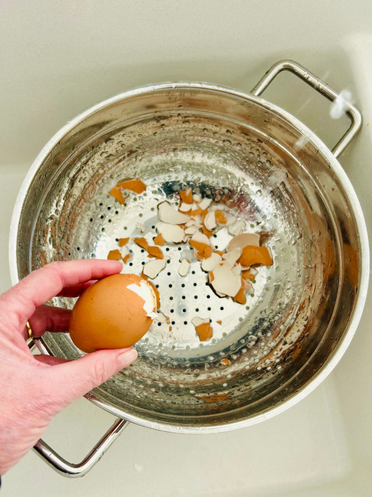 An egg is starting to have its shell peeled under running water with a colander below to catch the shells.