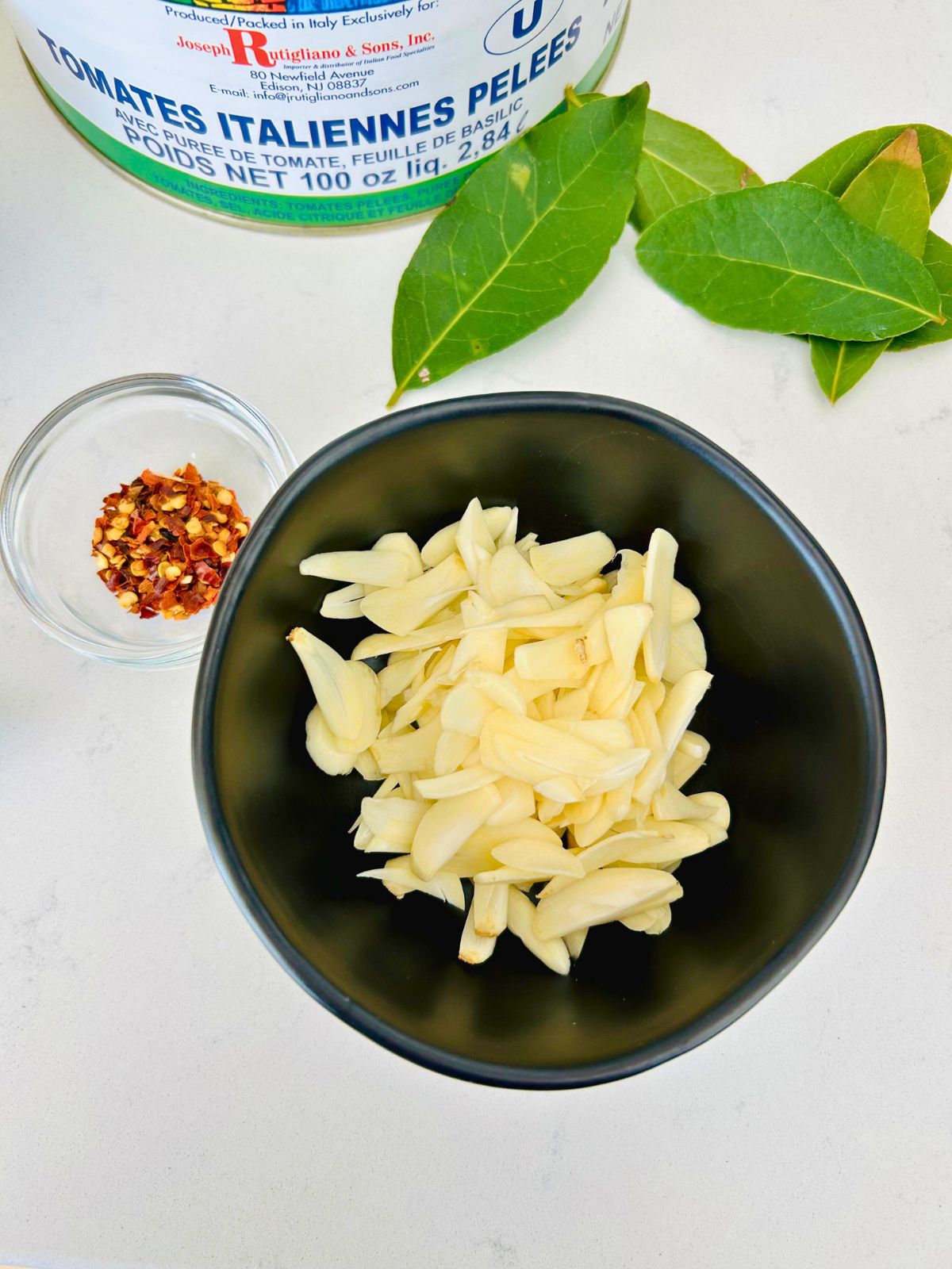 Sliced garlic prepped in a black bowl next to crushed red pepper and bay leaves.