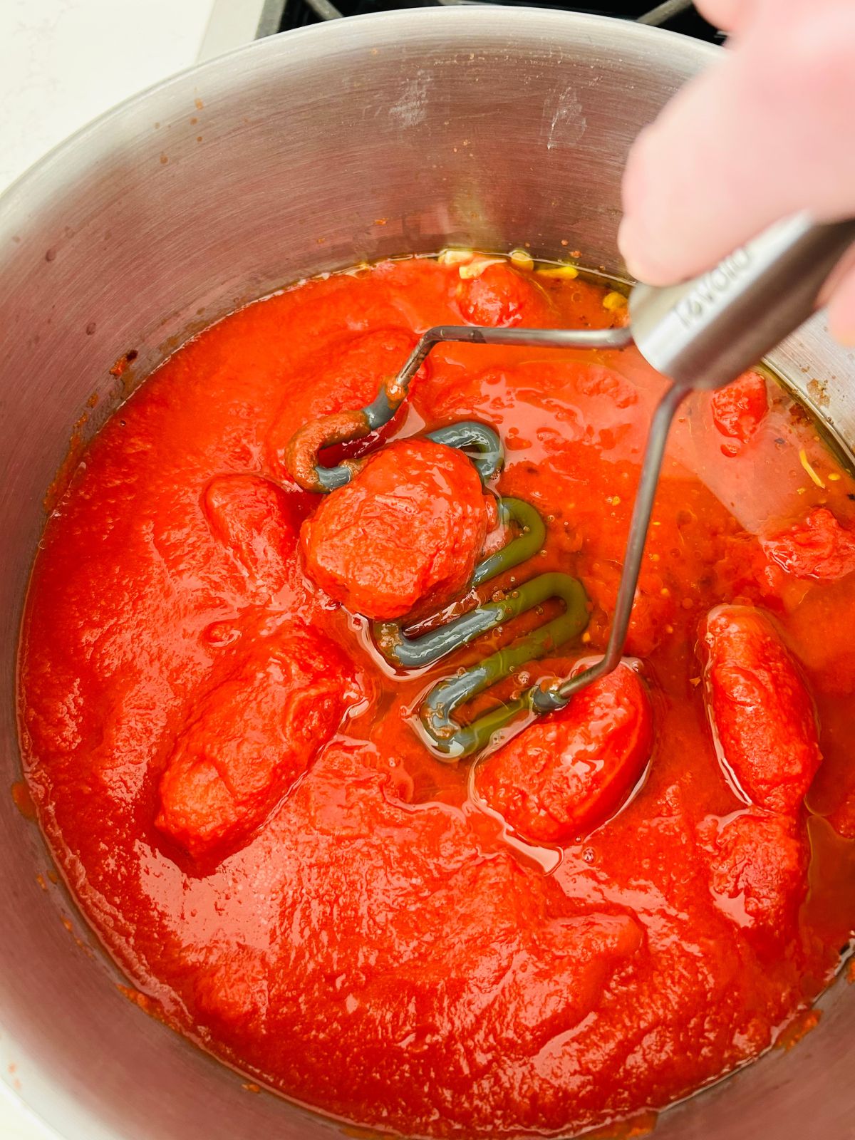 A potato masher is used to press down on the whole canned tomatoes so they become smaller pieces.