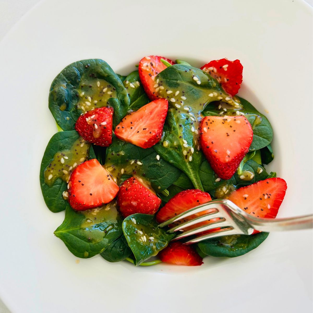 A fork stabbing some strawberry spinach salad with apple cider vinaigrette from a white bowl.