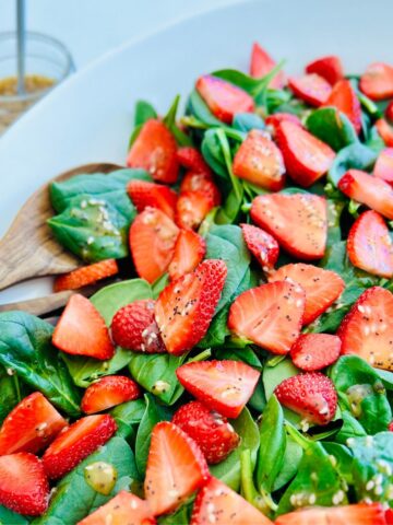 A bowl of strawberry spinach salad with salad tongs, and a jar or apple cider vinaigrette.