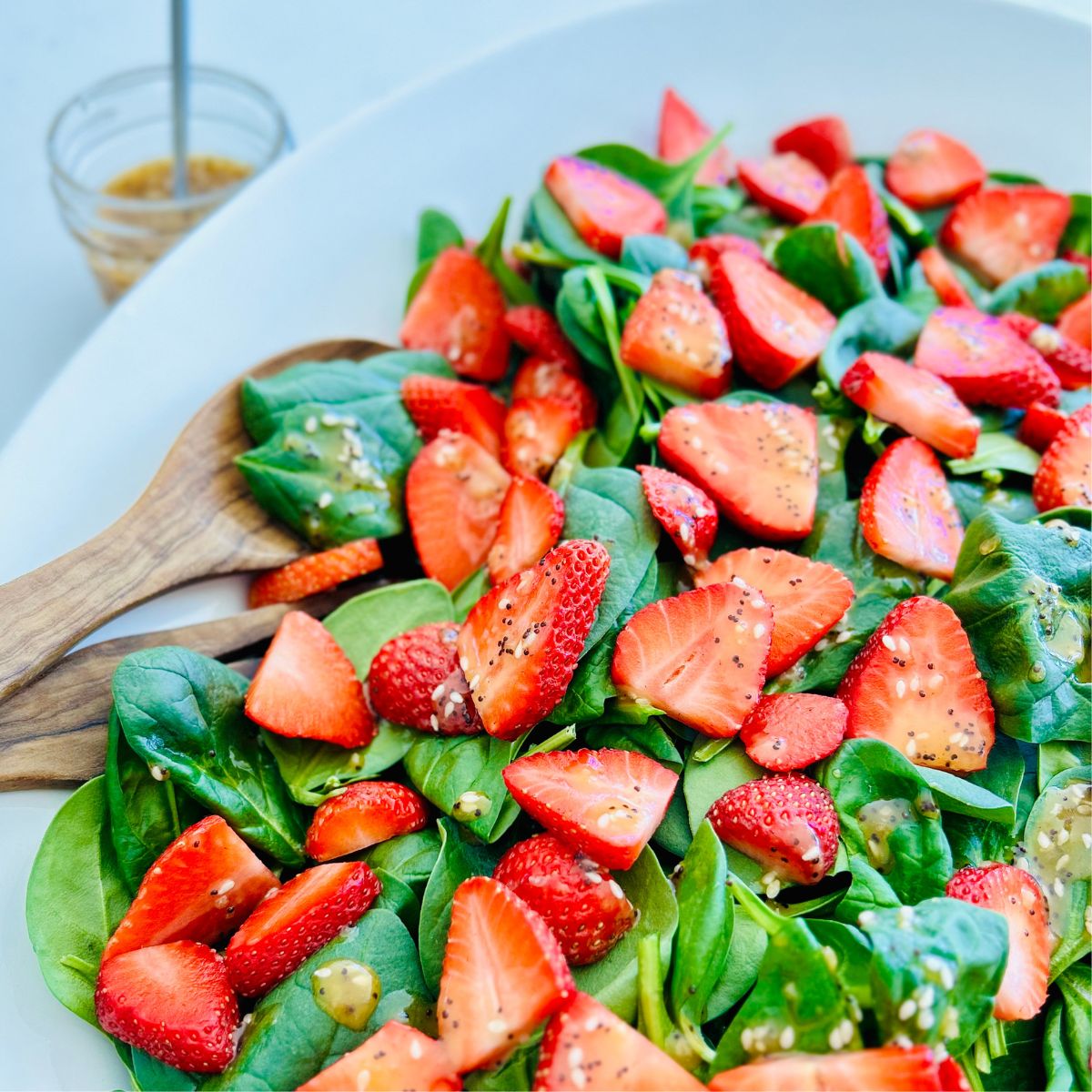 A bowl of strawberry spinach salad with salad tongs, and a jar or apple cider vinaigrette.