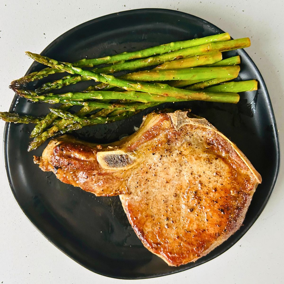 A pan-seared bone-in pork chop with sauteed asparagus on a black plate.