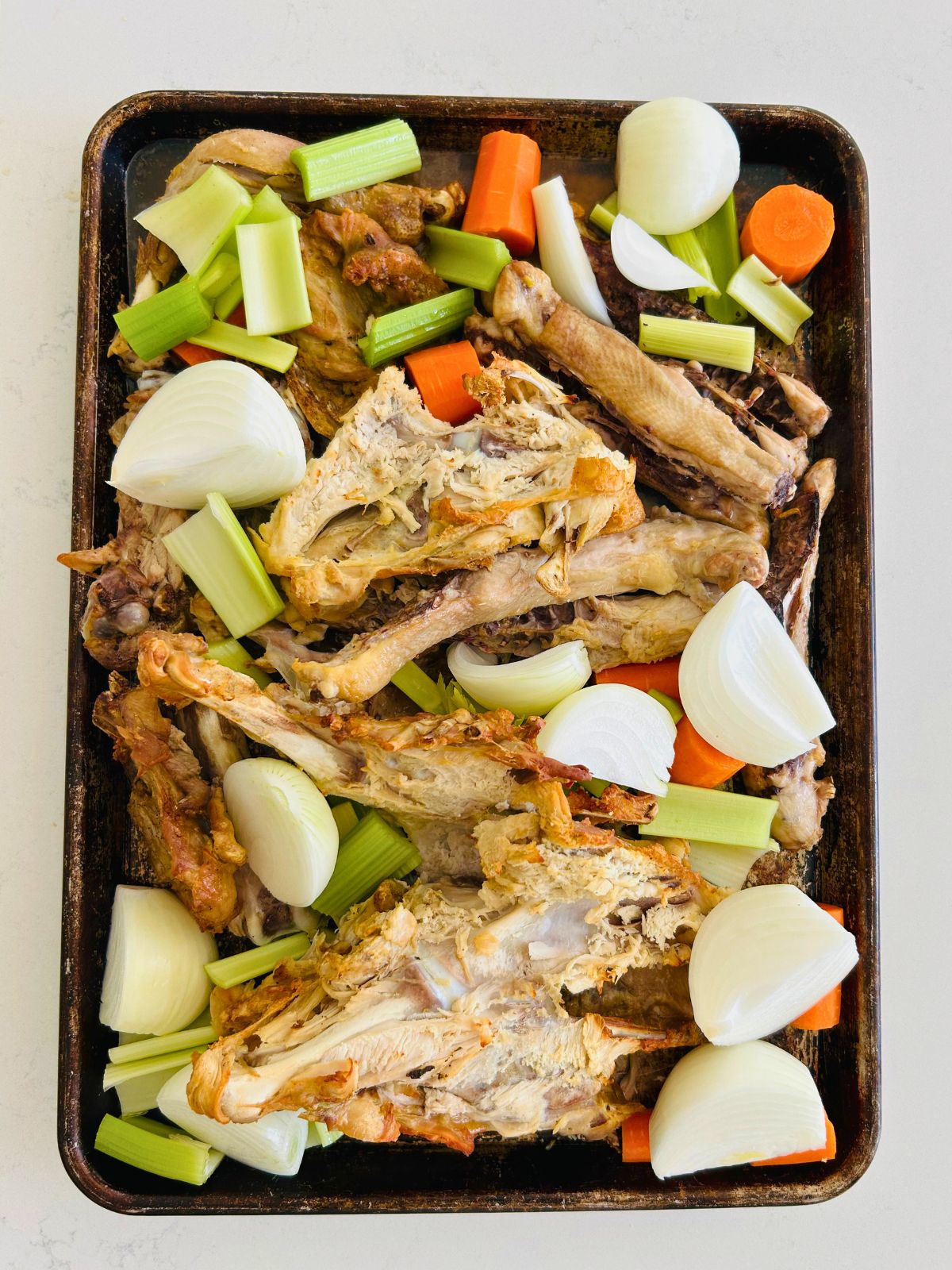 Partially roasted chicken frames and chunks of onion, celery, and carrots on a cookie sheet.