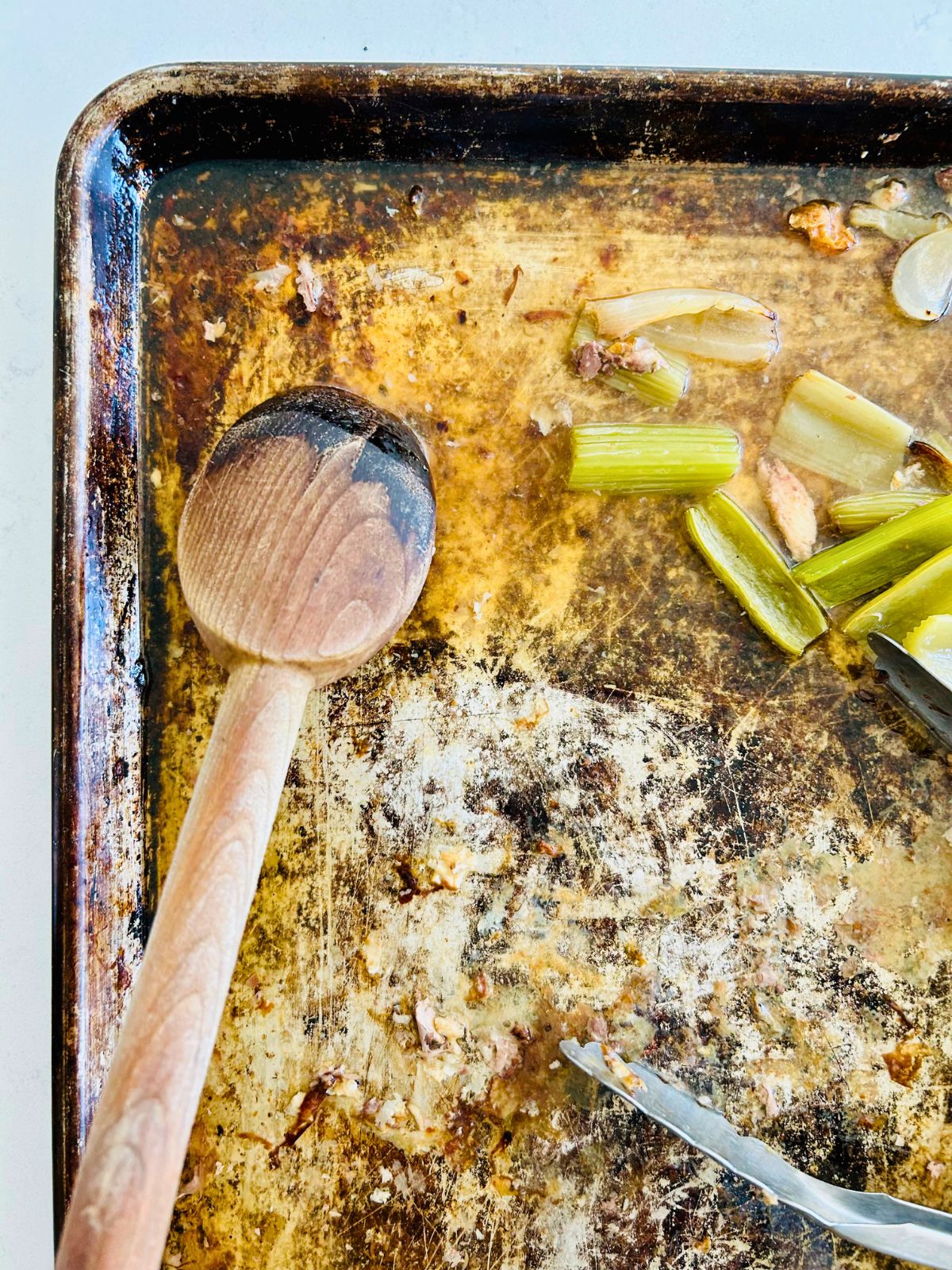 A wooden spoon is scraping the bits from the empty sheet pan after roasting the bones and mirepoix.