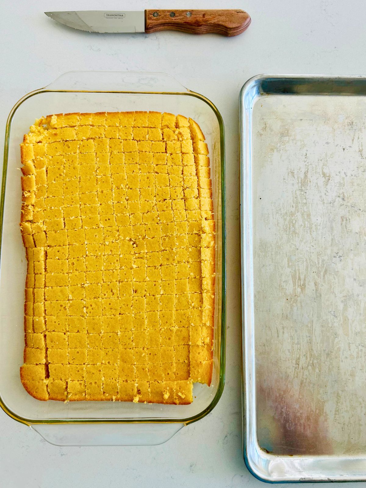 Cooked cornbread in a pyrex casserole that has been cubed with a steak knife next to a cookie sheet.