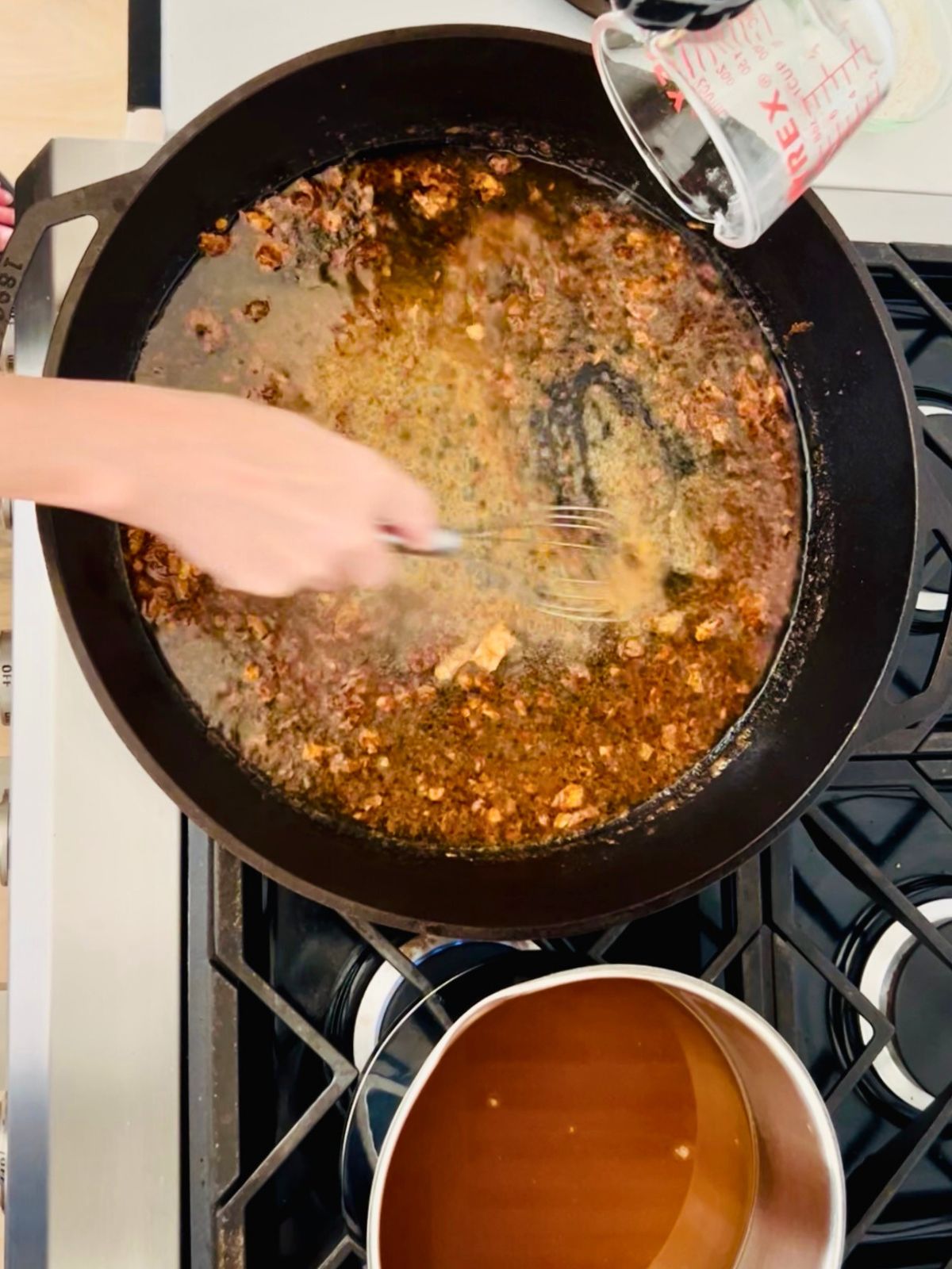 White wine is whisked into the turkey drippings of the cast iron skillet with a pot of warm brown chicken stock next to it.