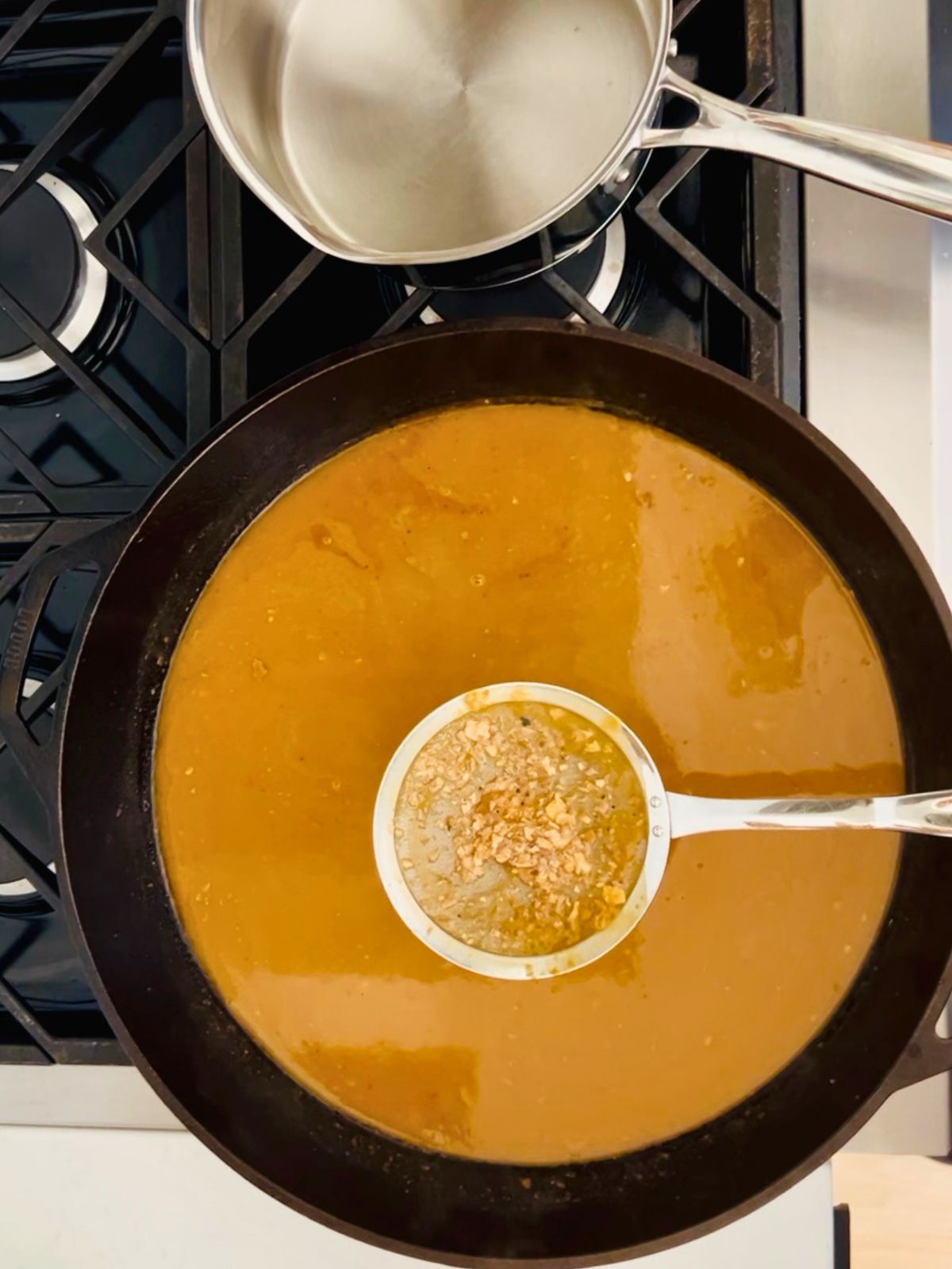 A fine mesh skimmer spoon is filtering the solids from the simmering turkey gravy in a large cast iron skillet.