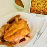 Whole roasted turkey in a white platter with pan gravy and stuffing casserole next to it.