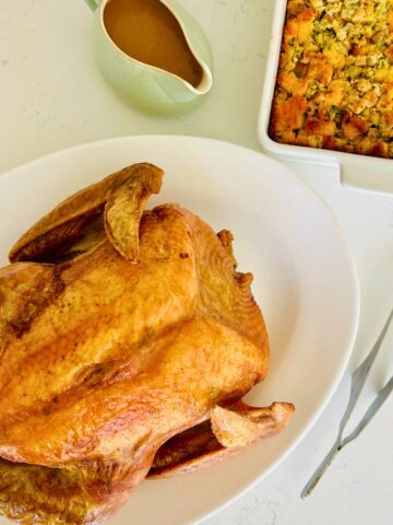 Whole roasted turkey in a white platter with pan gravy and stuffing casserole next to it.