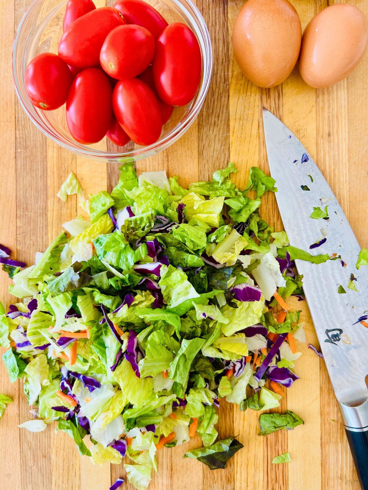 Chopped lettuce on a cutting board next to a bowl of tomatoes and eggs.