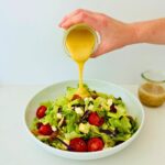 Honey mustard is dripping into a bowl of Houston's traditional salad with a cup of hot bacon dressing next to it.