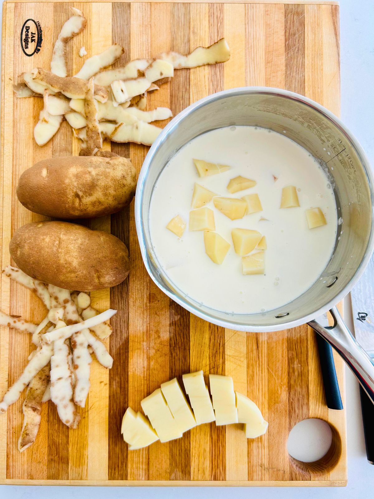 A pot of cream with cubed potatoes in it on a wooden cutting board with cubed and whole potatoes.