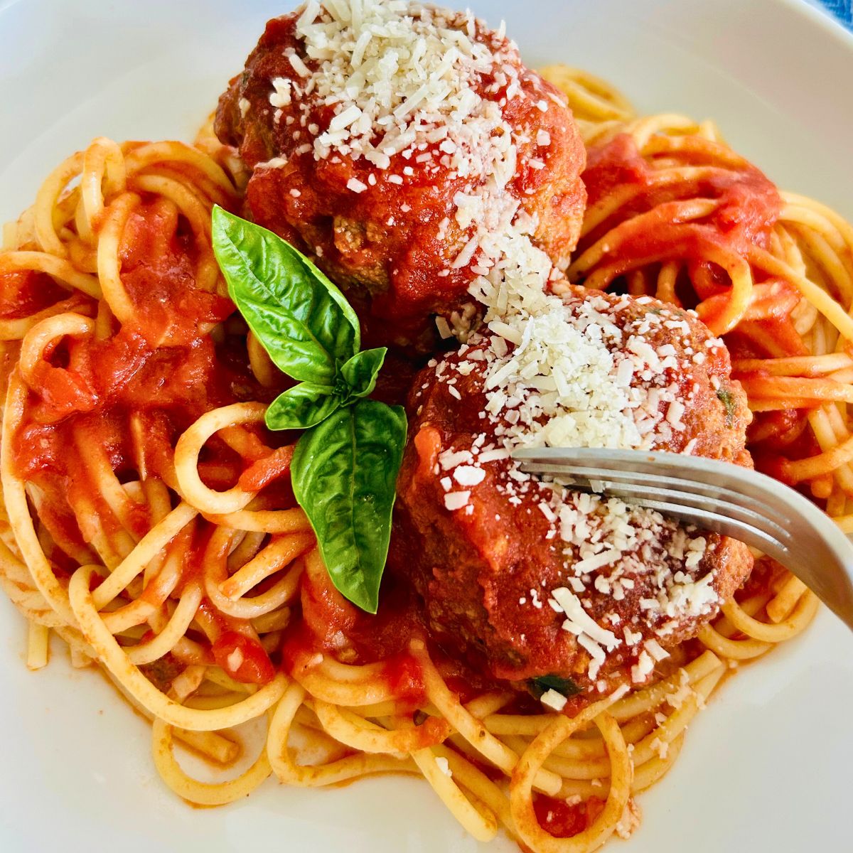A fork is splitting a meatball sitting on top of spaghetti noodles with parmesan cheese and fresh basil on top.