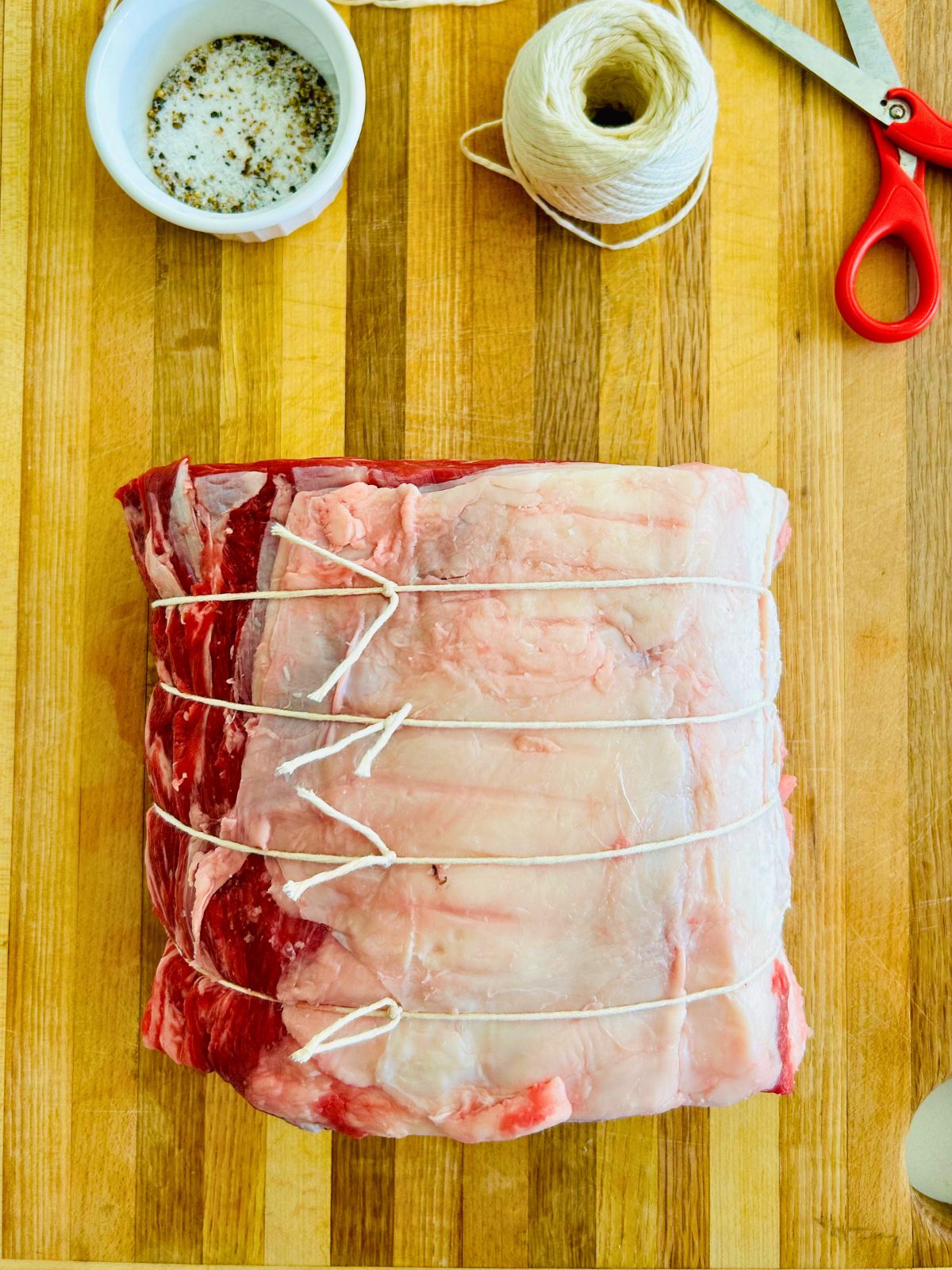 Prime rib roast tied with butcher twine next to a bowl of salt and pepper on a cutting board.