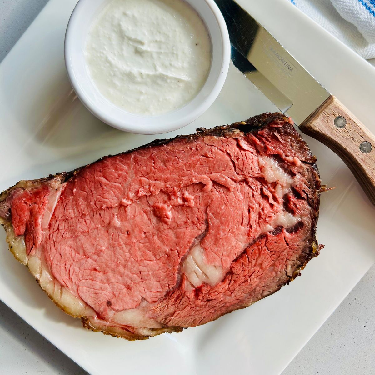 A slice of slow roasted prime rib on a white plate next to horseradish sauce and a steak knife.