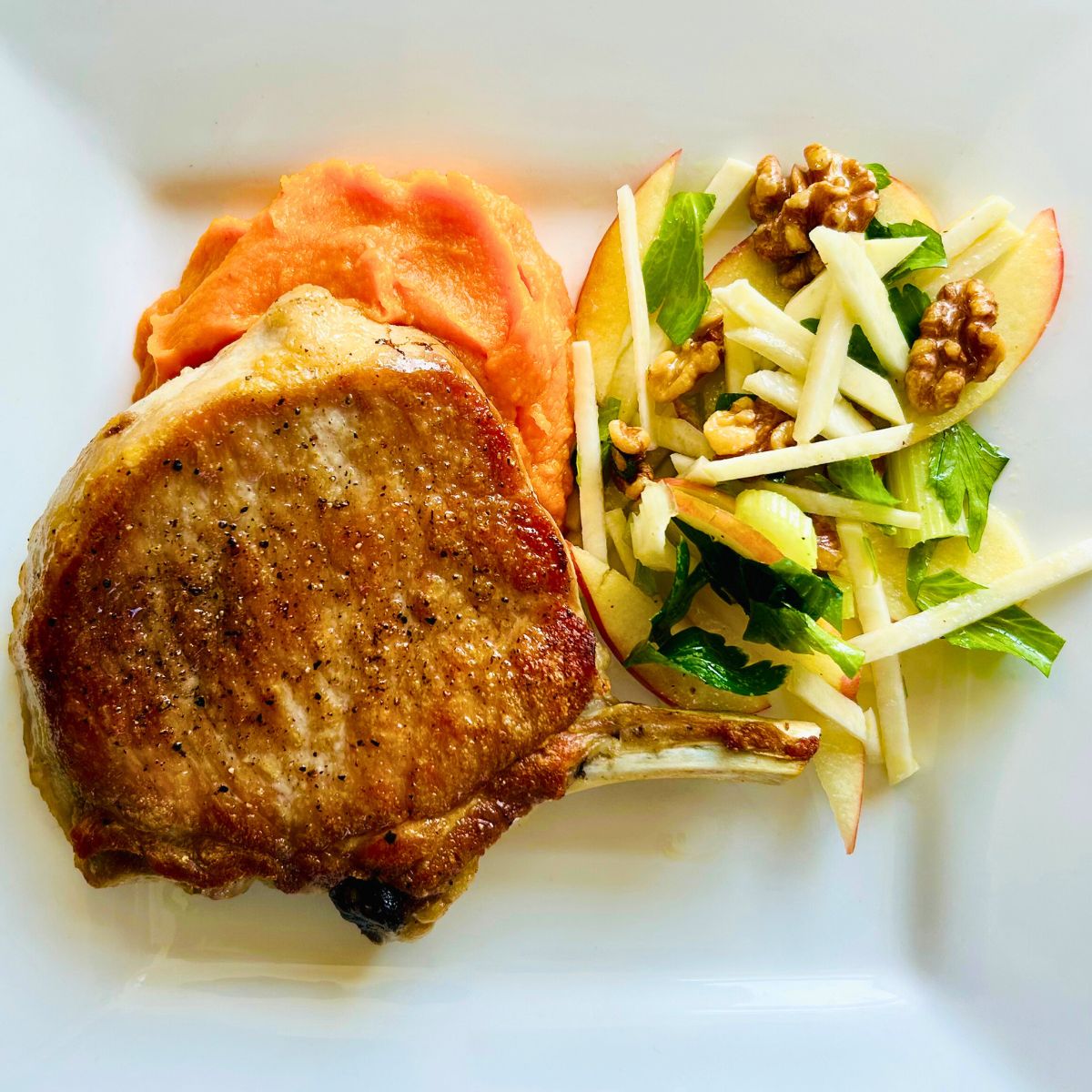 A pan seared pork chop on top of chipotle sweet potatoes next to apple kohlrabi salad on a white plate.
