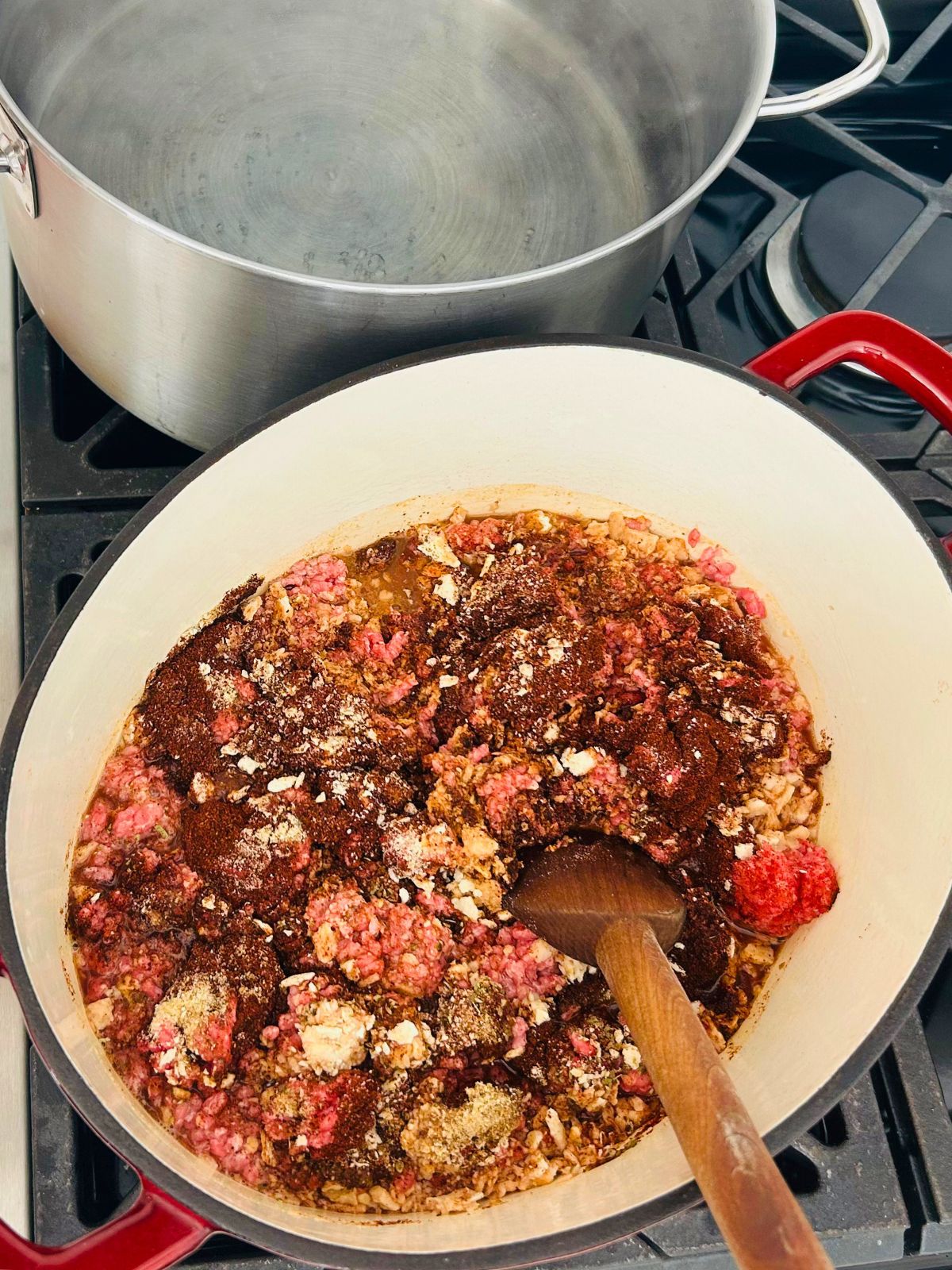 A Dutch oven containing ground beef, chili spices, crushed saltines, and a wooden spoon is on a stovetop.