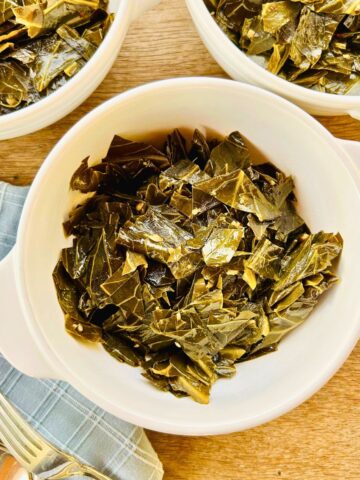 Three white bowls containing braised collard greens next to forks and blue napkins.