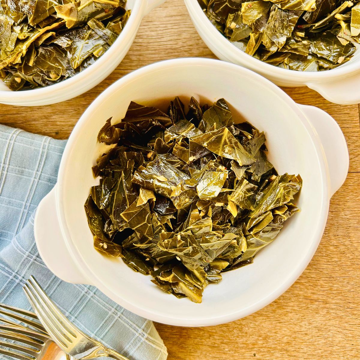 Three white bowls containing braised collard greens next to forks and blue napkins.