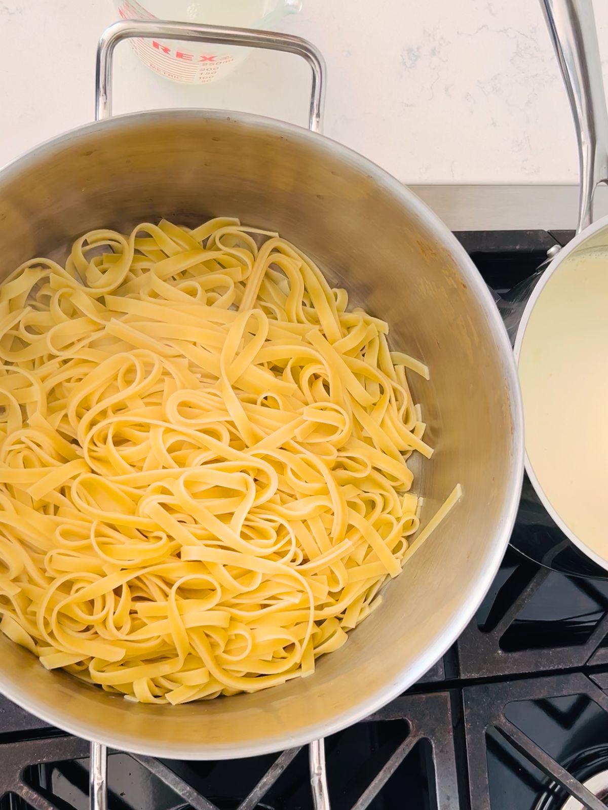 Fettuccine pasta cooked and drained in a pot next to a smaller pot of creamy Alfredo sauce.