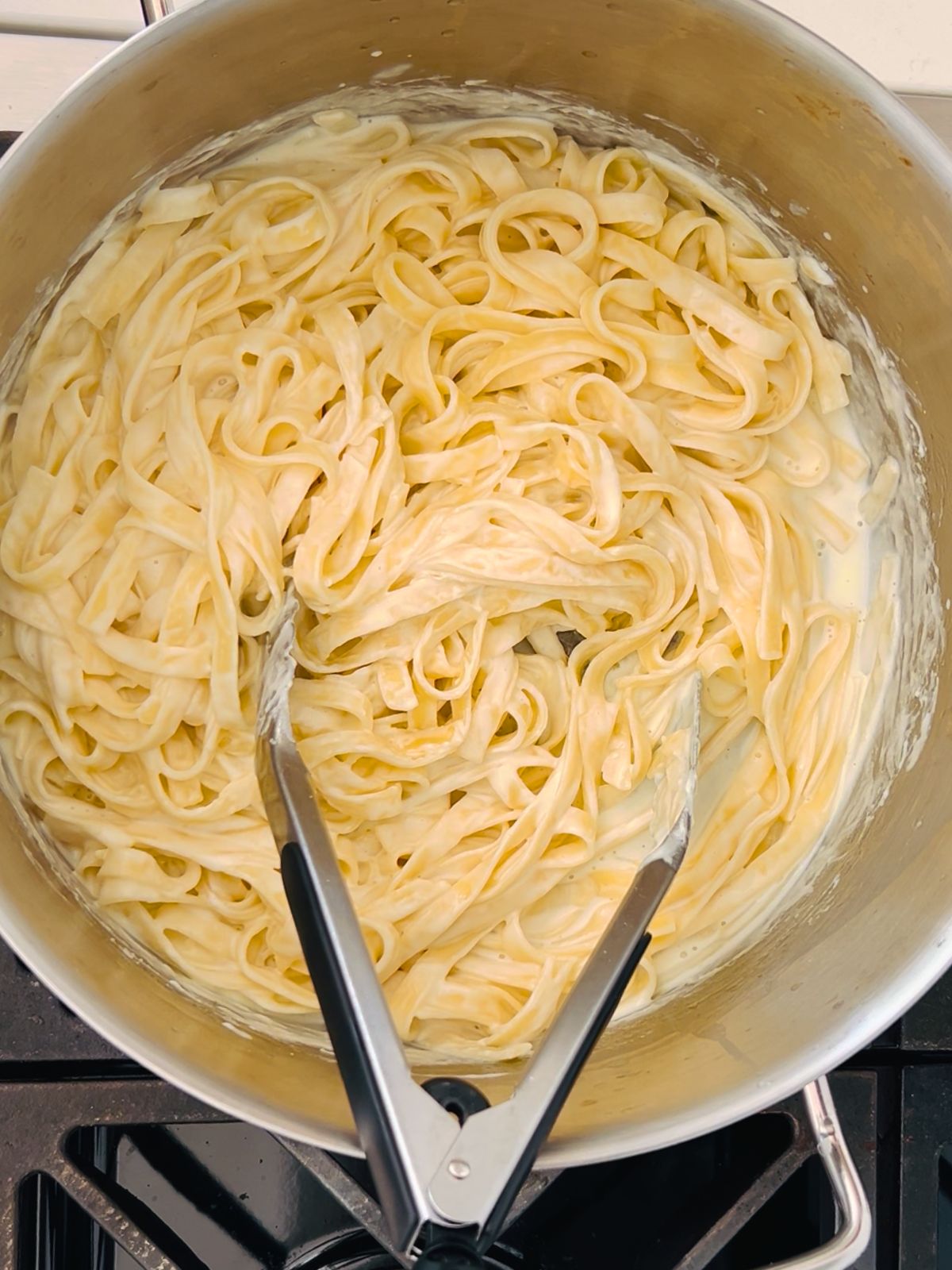 Fettuccine noodles sauced with Alfredo sauce in a pot with tongs resting in it.