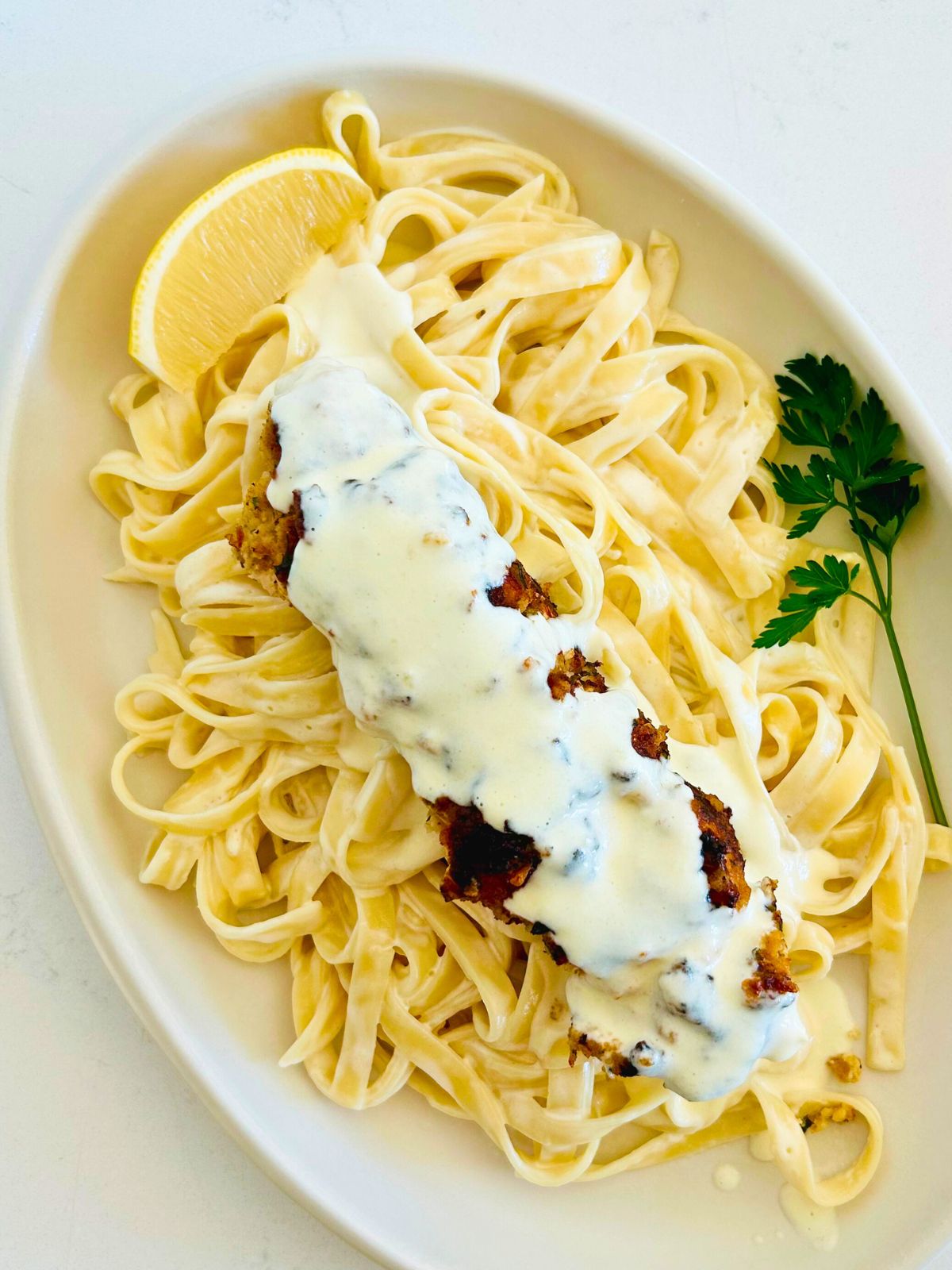 A white oval plate with fettuccini Alfredo topped with chicken spiedini covered with more Alfredo sauce.