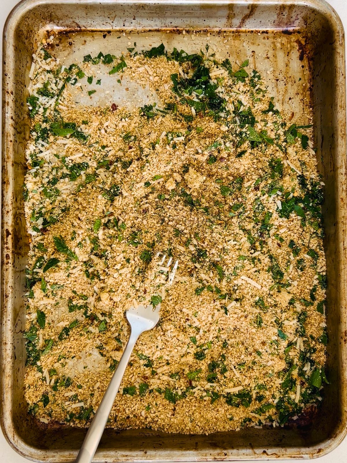Seasoned Italian breadcrumbs, Pecorino Romano cheese, chopped garlic, and parsley are combined by a fork in a cookie sheet.