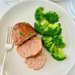 An individual ham loaf has been sliced open and nestled next to steam broccoli on a white plate.