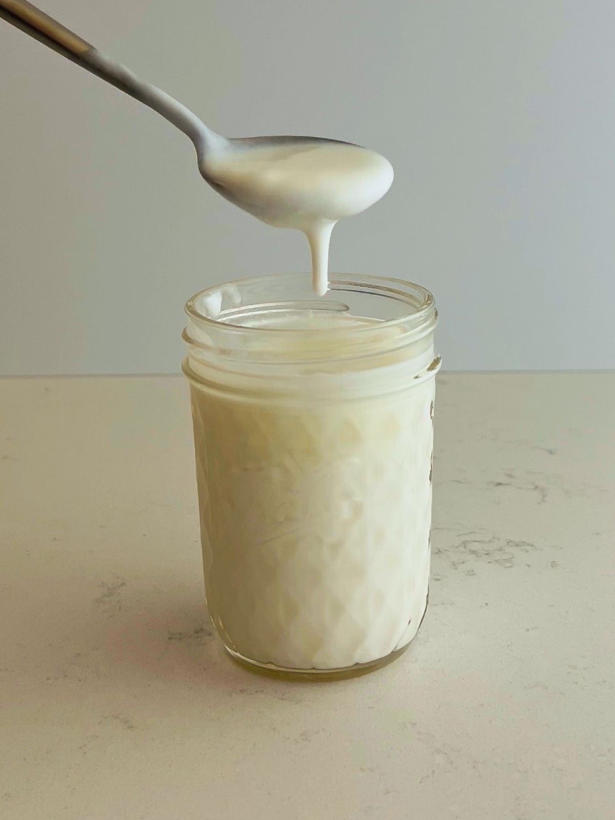 A spoon with thick and drippy cultured buttermilk is held over the mason jar on the kitchen counter.