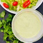 A jar of green goddess salad dressing next to a bowl of salad and parsley sprigs.