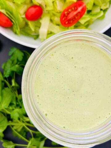 A jar of green goddess salad dressing next to a bowl of salad and parsley sprigs.