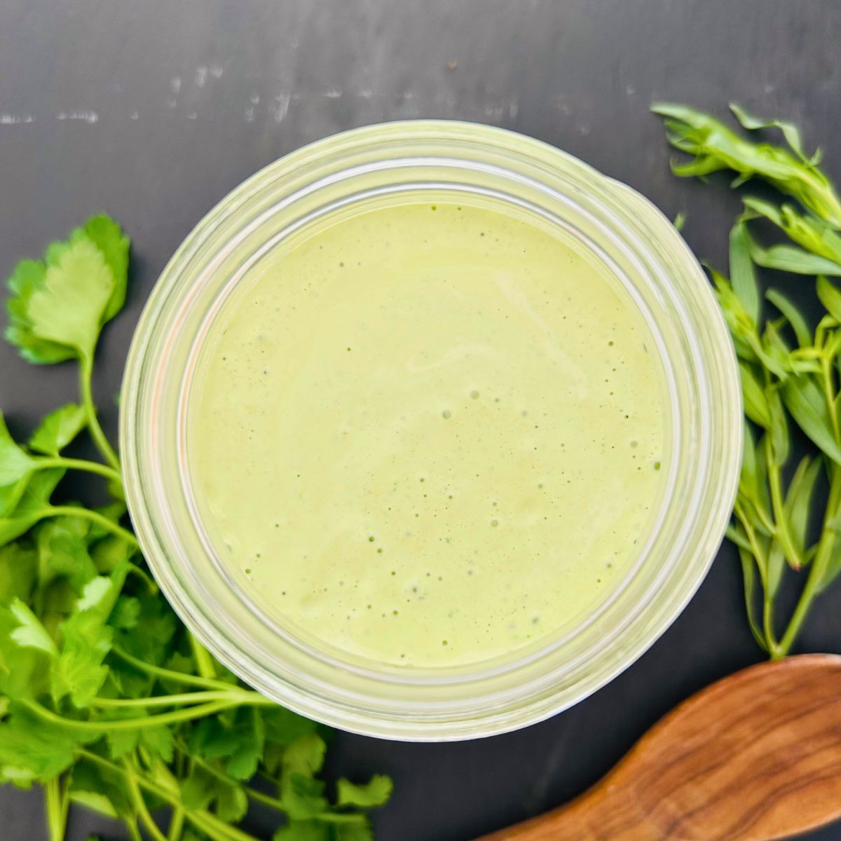 A glass jar of green goddess salad dressing recipe next to fresh tarragon, parsley, and a wooden spoon.