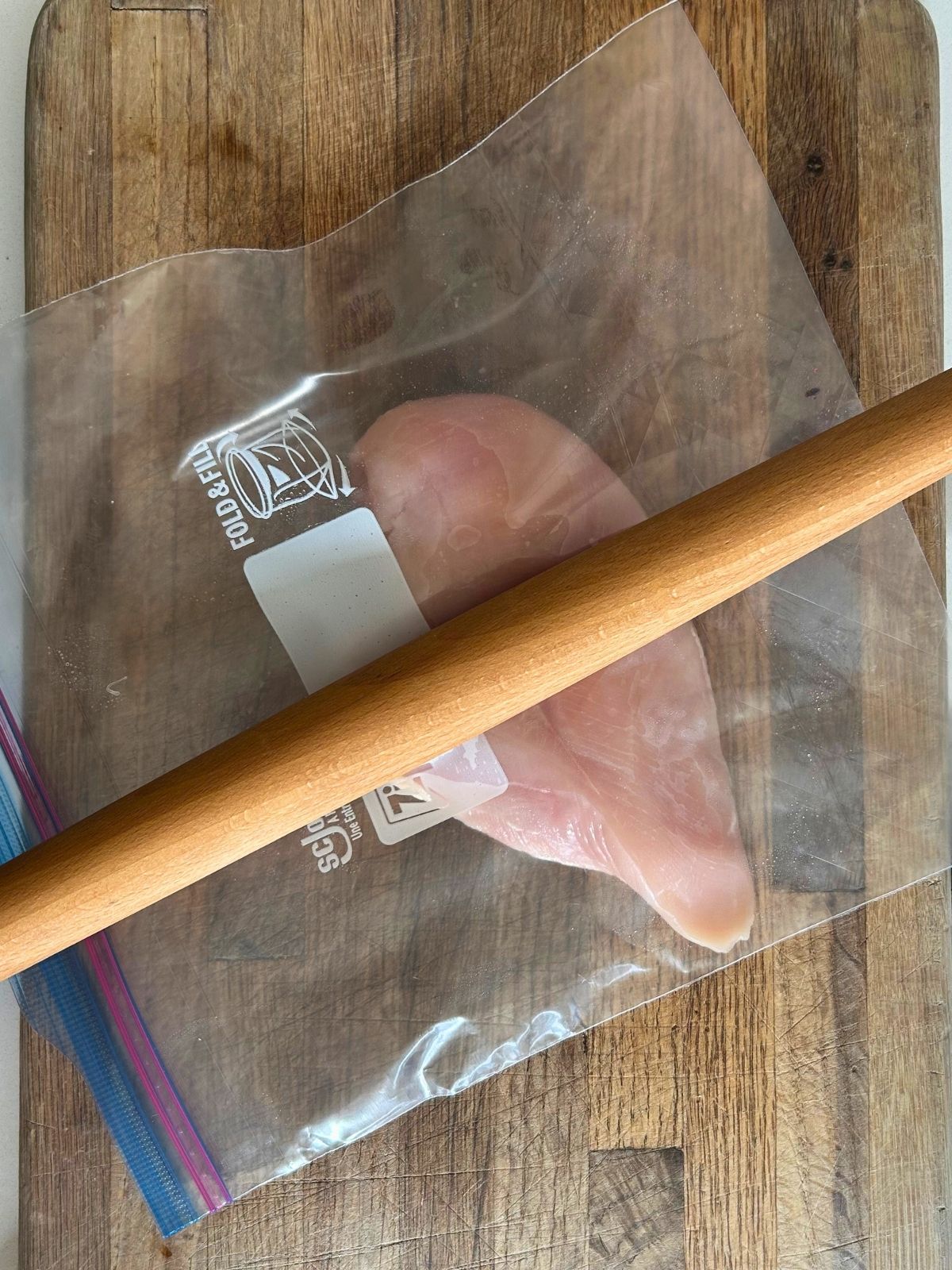A rolling pin is hitting the thick area of a chicken breast in a zip-lock bag to achieve an even thickness.