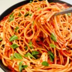 Tongs are pulling strands of bucatini mixed with tomatoes, fresh basil, and melted mozzarella in a skillet.