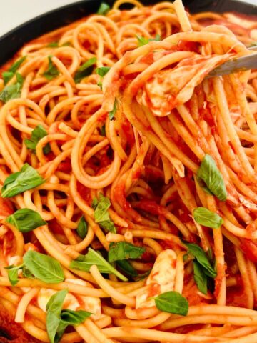 Tongs are pulling strands of bucatini mixed with tomatoes, fresh basil, and melted mozzarella in a skillet.