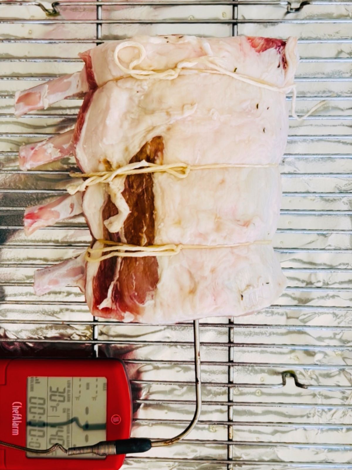Pork rib roast is tied and lying on a wire rack over a cookie sheet with a meat thermometer probe inserted.