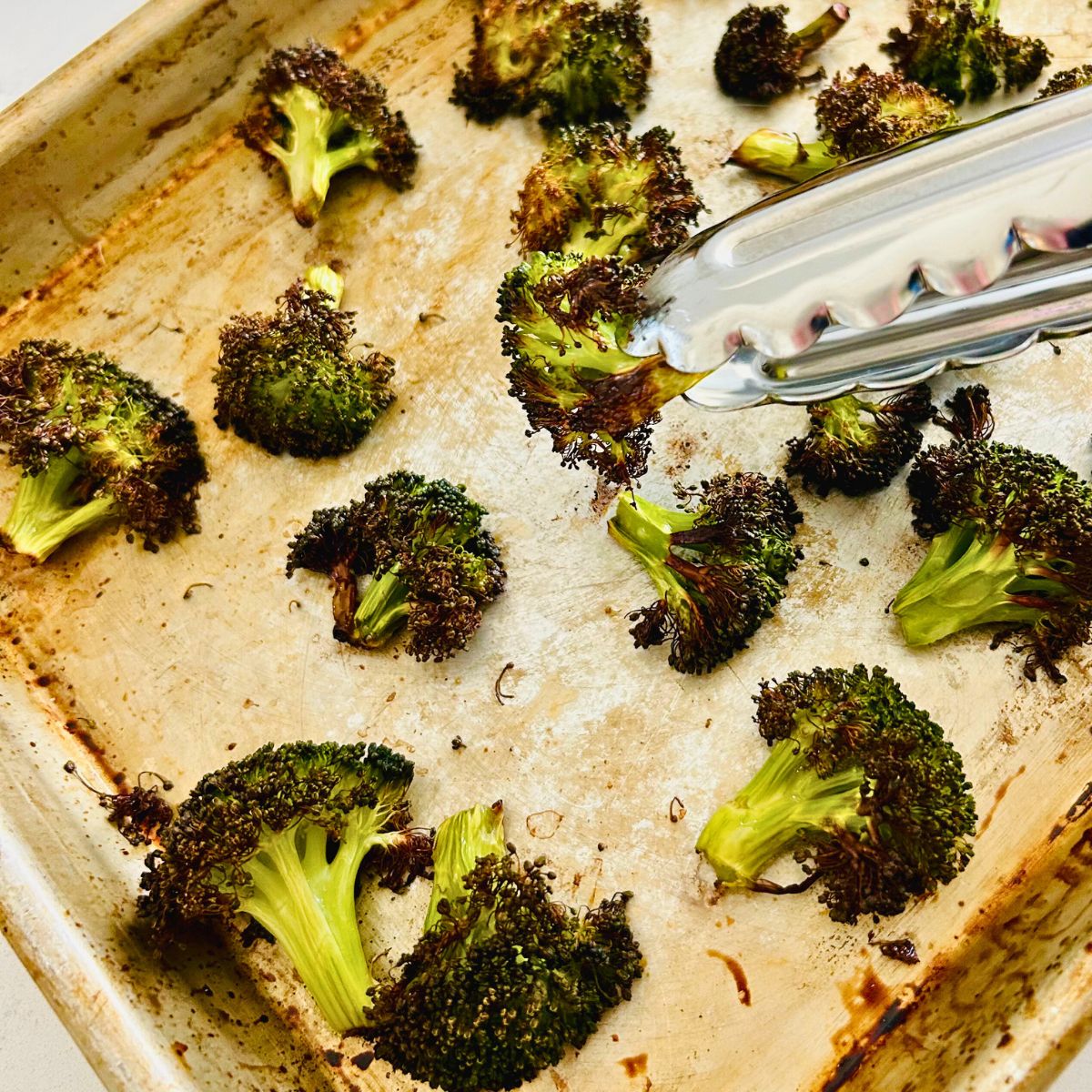 Tongs are holding a floret over a sheet pan of crispy roasted broccoli.