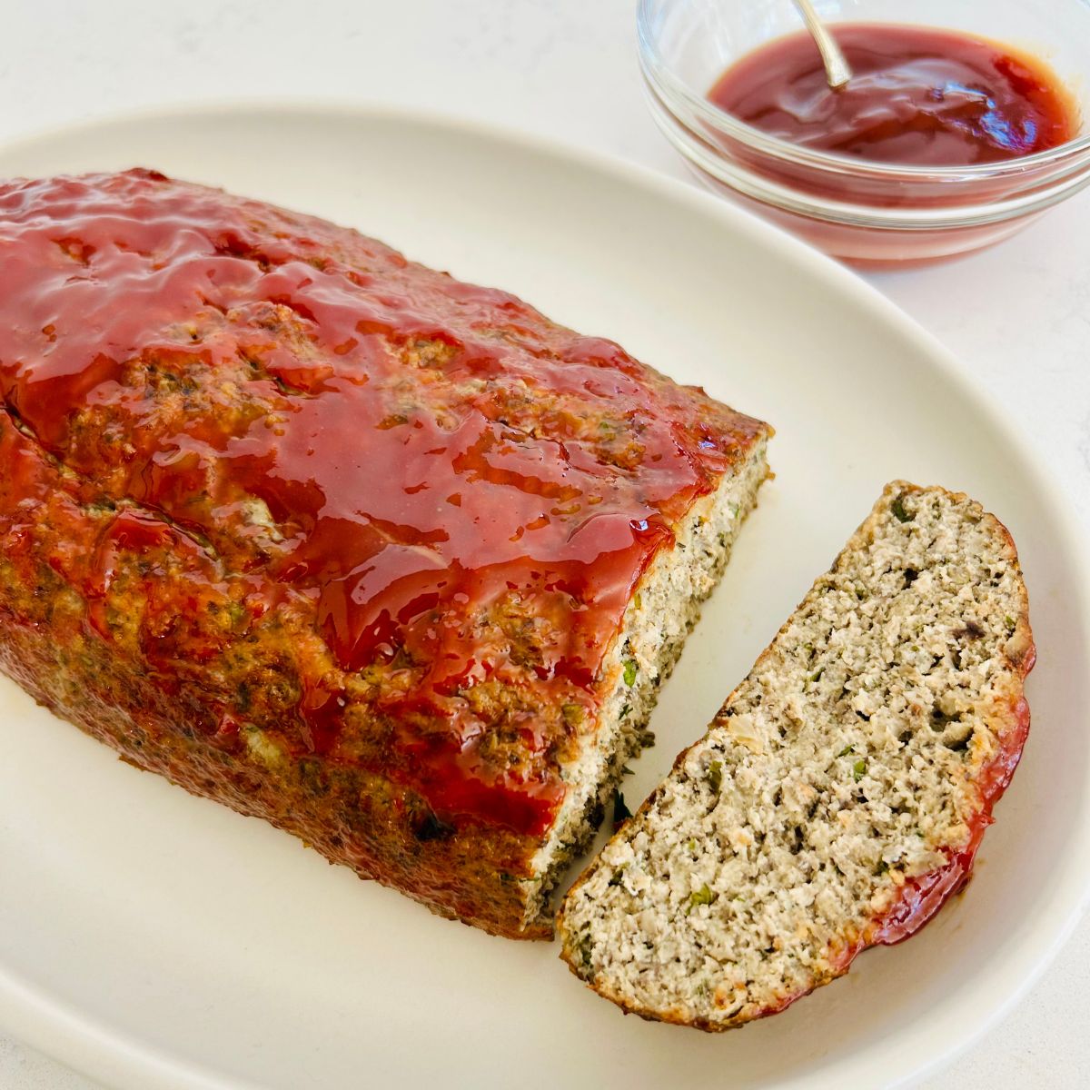 Turkey meatloaf with a sweet tangy glaze sliced on a white plate next to a bowl of extra glaze.