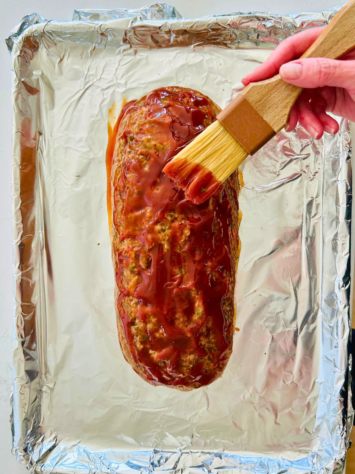 The sweet tangy glaze is getting brushed onto the formed turkey meatloaf sitting on a foil-lined baking sheet.
