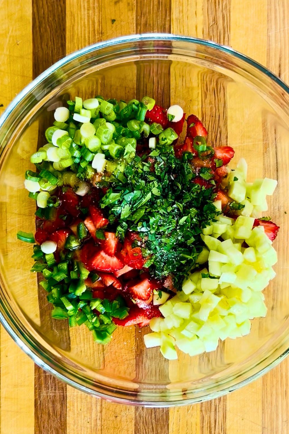 Chopped strawberries, cucumber, jalapeno, cilantro, and green onions in a clear glass mixing bowl.
