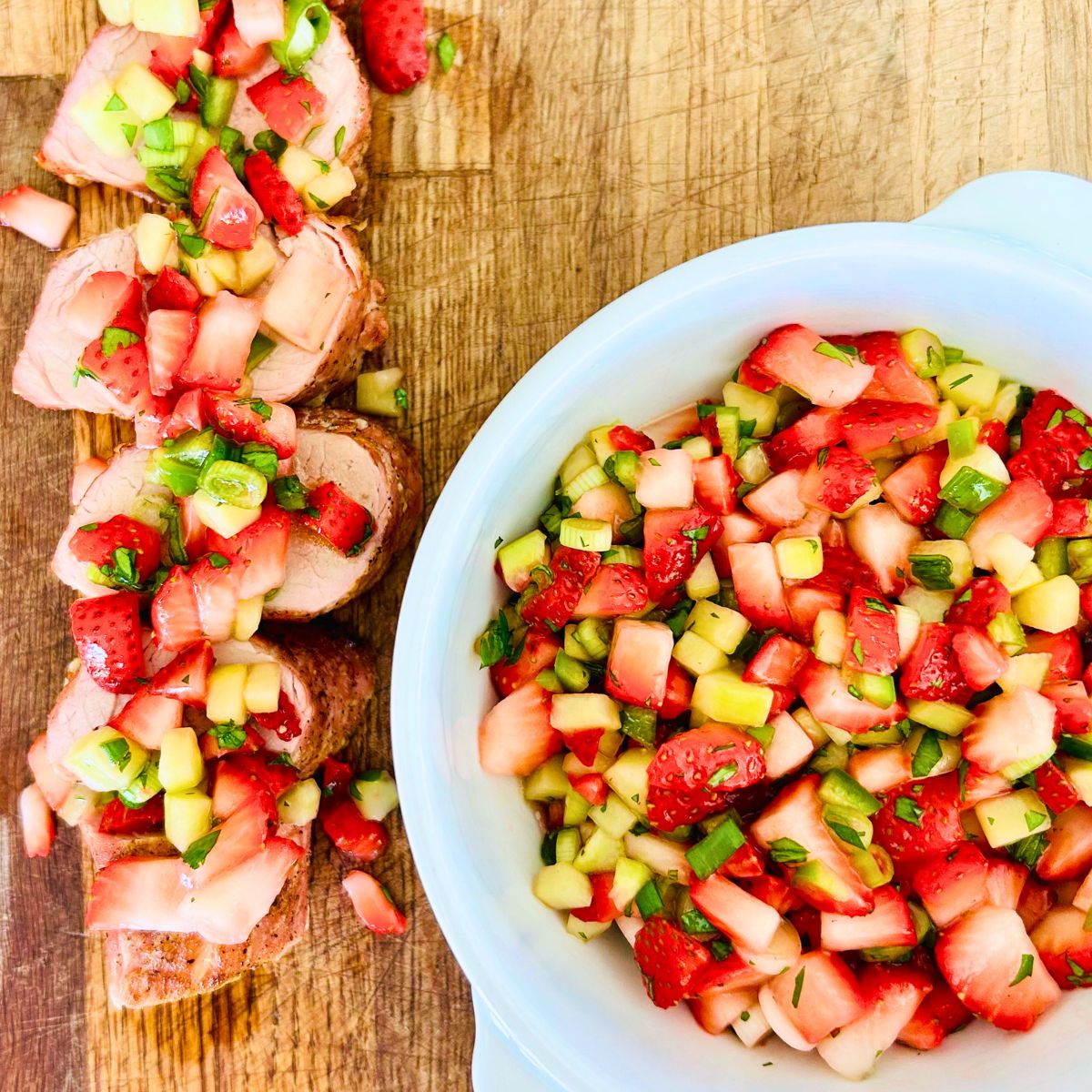 Strawberry salsa recipe in a white bowl and sprinkled over a row of sliced, grilled pork tenderloin.