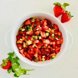 Strawberry salsa recipe in a white bowl next to whole strawberries and cilantro sprigs.