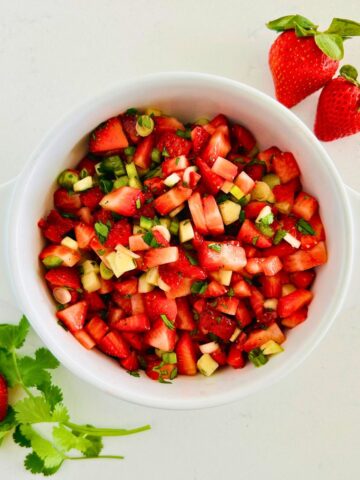Strawberry salsa recipe in a white bowl next to whole strawberries and cilantro sprigs.