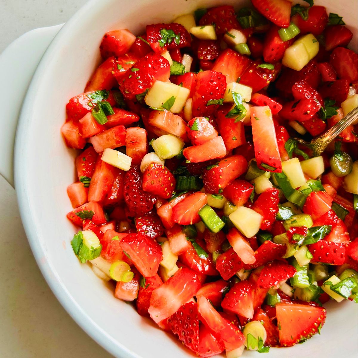 Chopped strawberries, cilantro, green onions, cucumbers, and jalapeno in a white bowl with a spoon resting inside.
