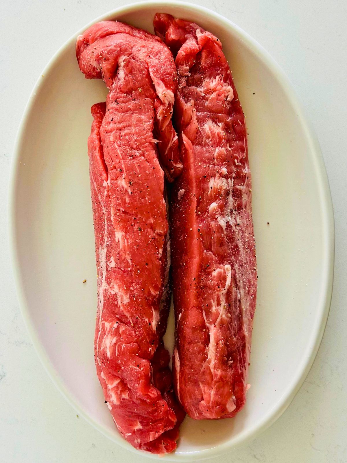Seasoned pork tenderloins ready to cook resting on a white oval platter.