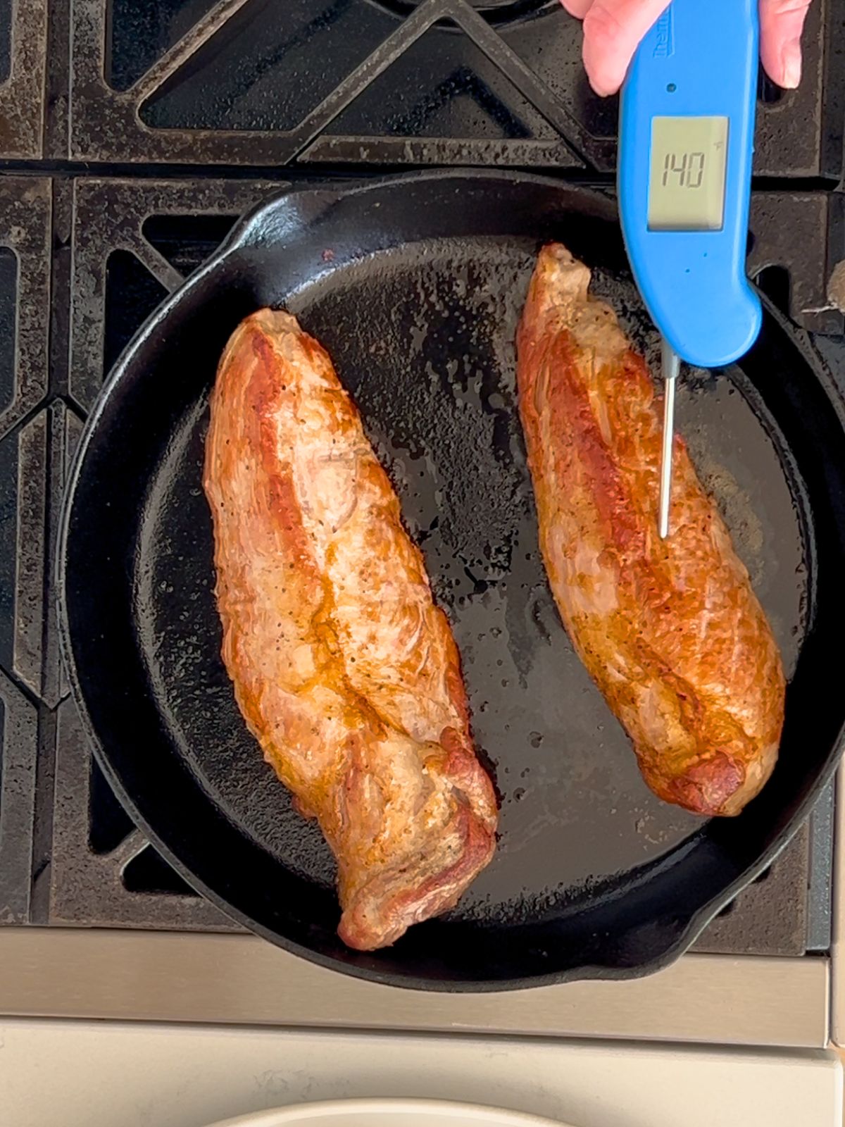Two roasted pork tenderloins in a cast iron skillet with a meat thermometer checking the internal temperature.
