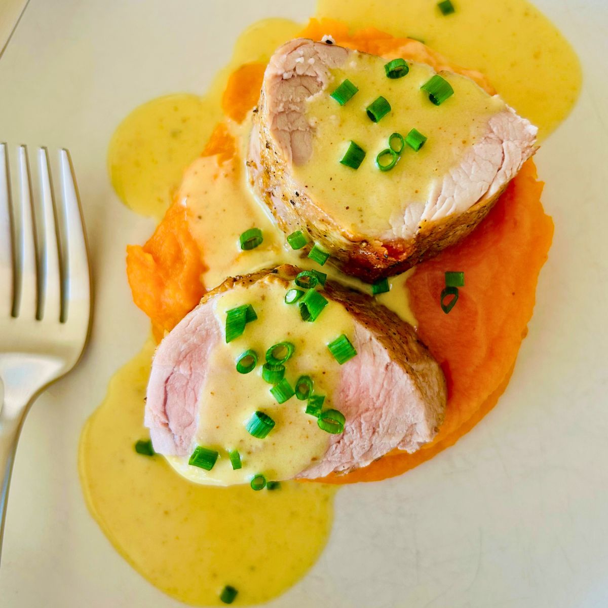 Pork tenderloin slices, curry butter, sweet potatoe puree and chives on a white plate with a fork to the side.